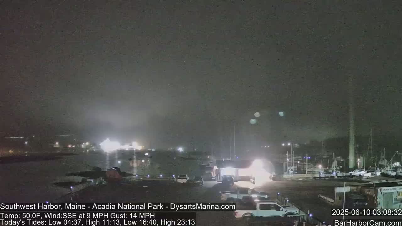 A nighttime harbor scene with hazy conditions and several boats docked, illuminated by lights from buildings and vehicles on shore.