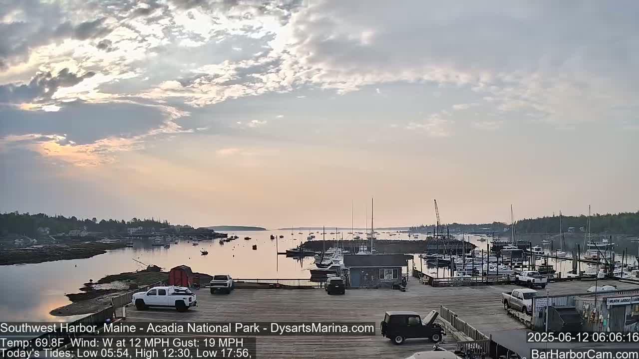 A harbor filled with boats at sunrise under a partly cloudy sky, with a gentle breeze and several vehicles parked near a dock.