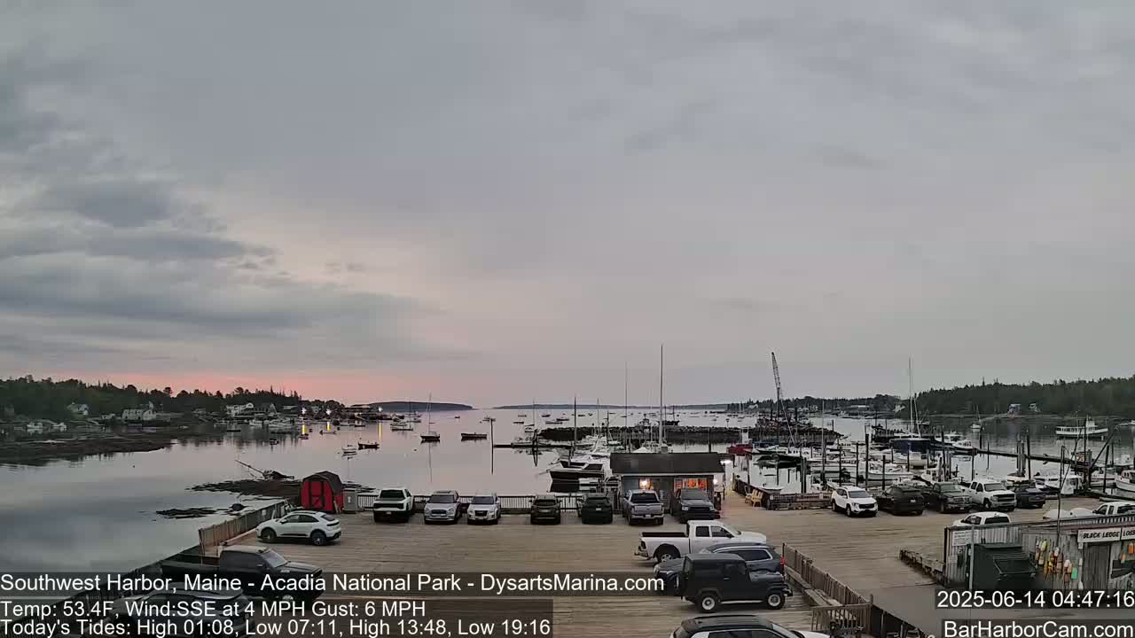 A calm harbor filled with boats is viewed from a parking lot under a mostly cloudy, dusky sky.
