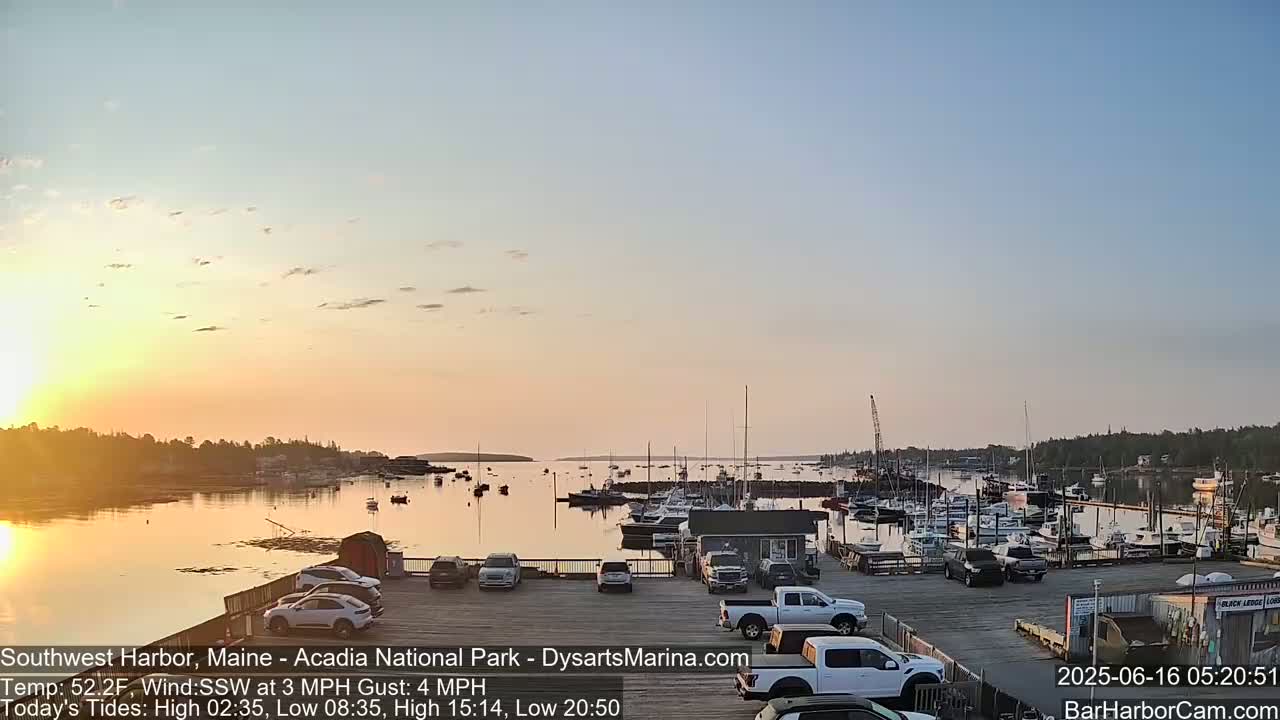 A calm harbor at sunset filled with numerous boats, cars parked in a lot beside it, and a calm body of water reflecting the sky.