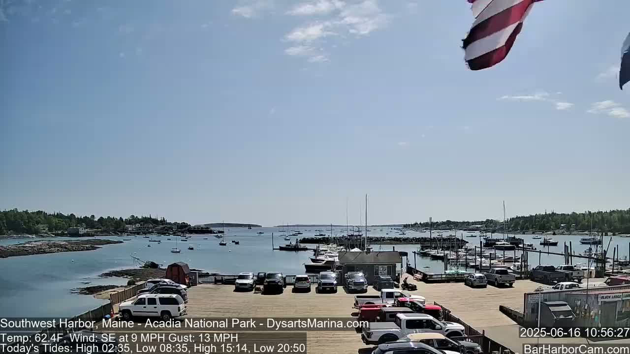 A harbor filled with boats under a mostly sunny sky, with a parking lot of cars in the foreground.