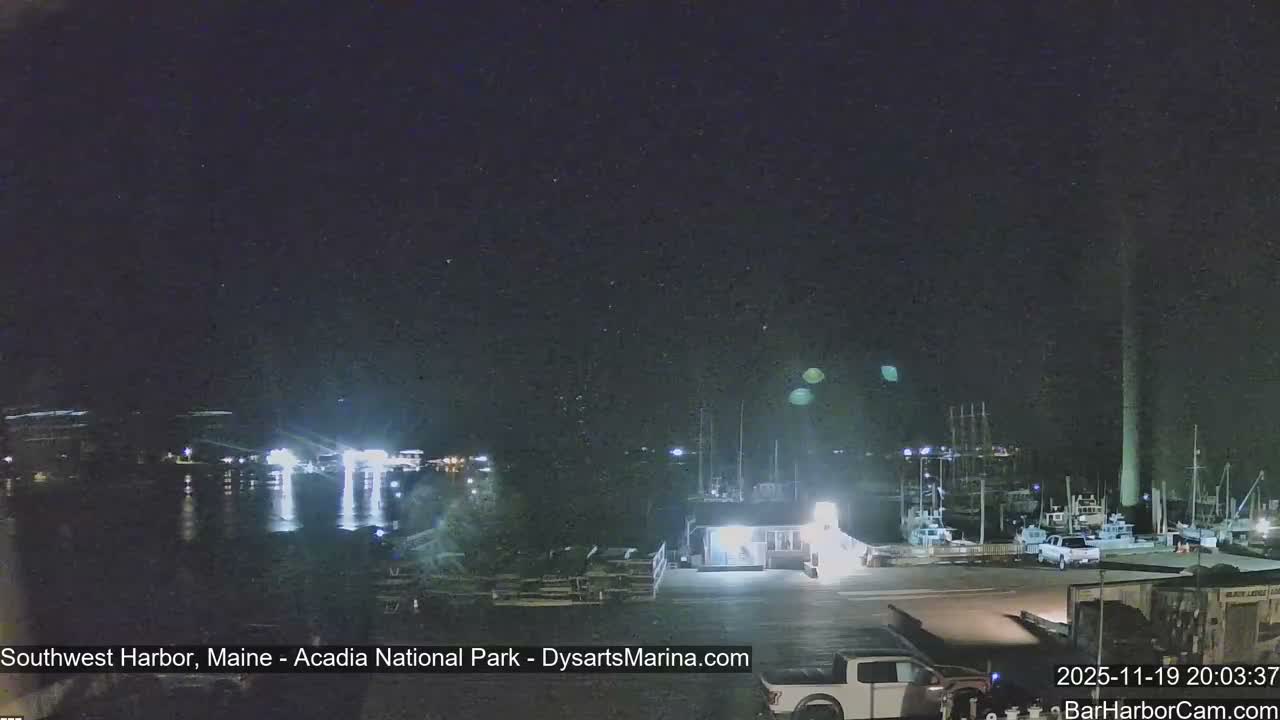 A clear night reveals a calm harbor scene illuminated by numerous waterfront lights reflecting on the water, with several boats docked and a large vertical structure on the right.