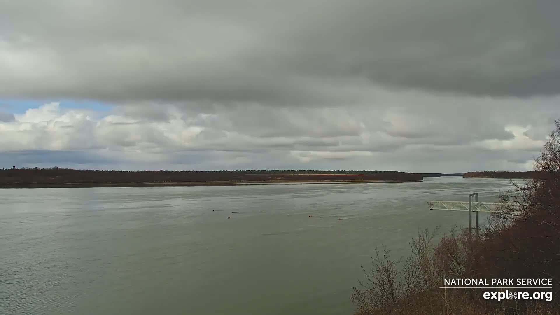A wide, calm river flows under an overcast sky, with a few small objects visible in the water and a metal structure extending into the river from the right.
