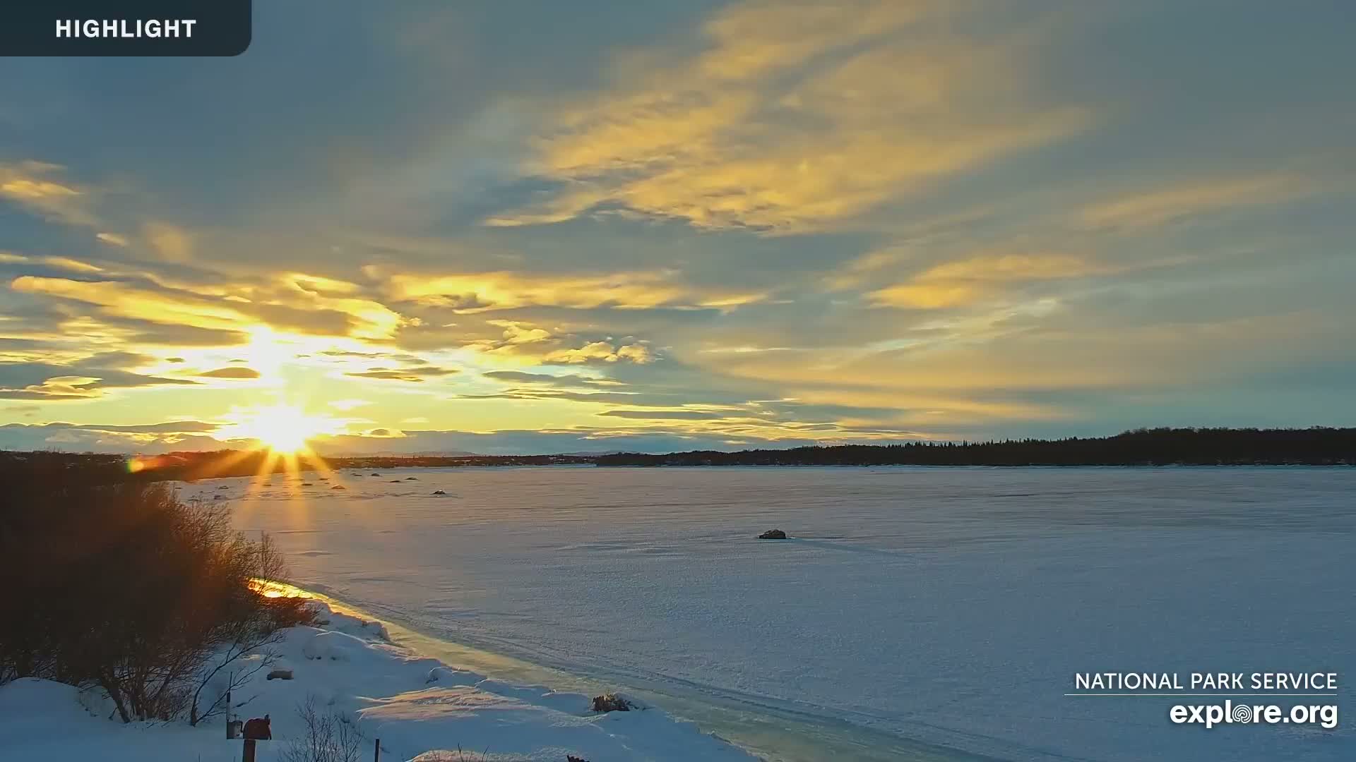 A vibrant sunset casts golden hues across a snow-covered, frozen lake, with a treeline visible in the background.