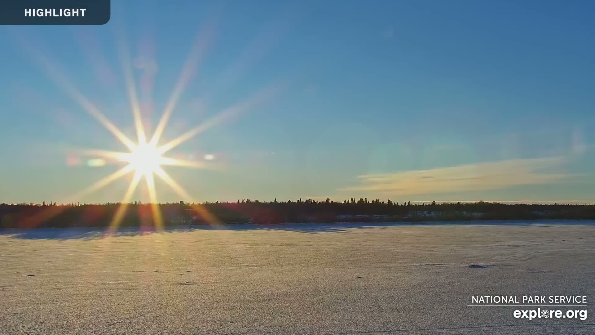 The bright sun shines over a frozen, snow-covered lake with a dark treeline in the background under a clear blue sky.