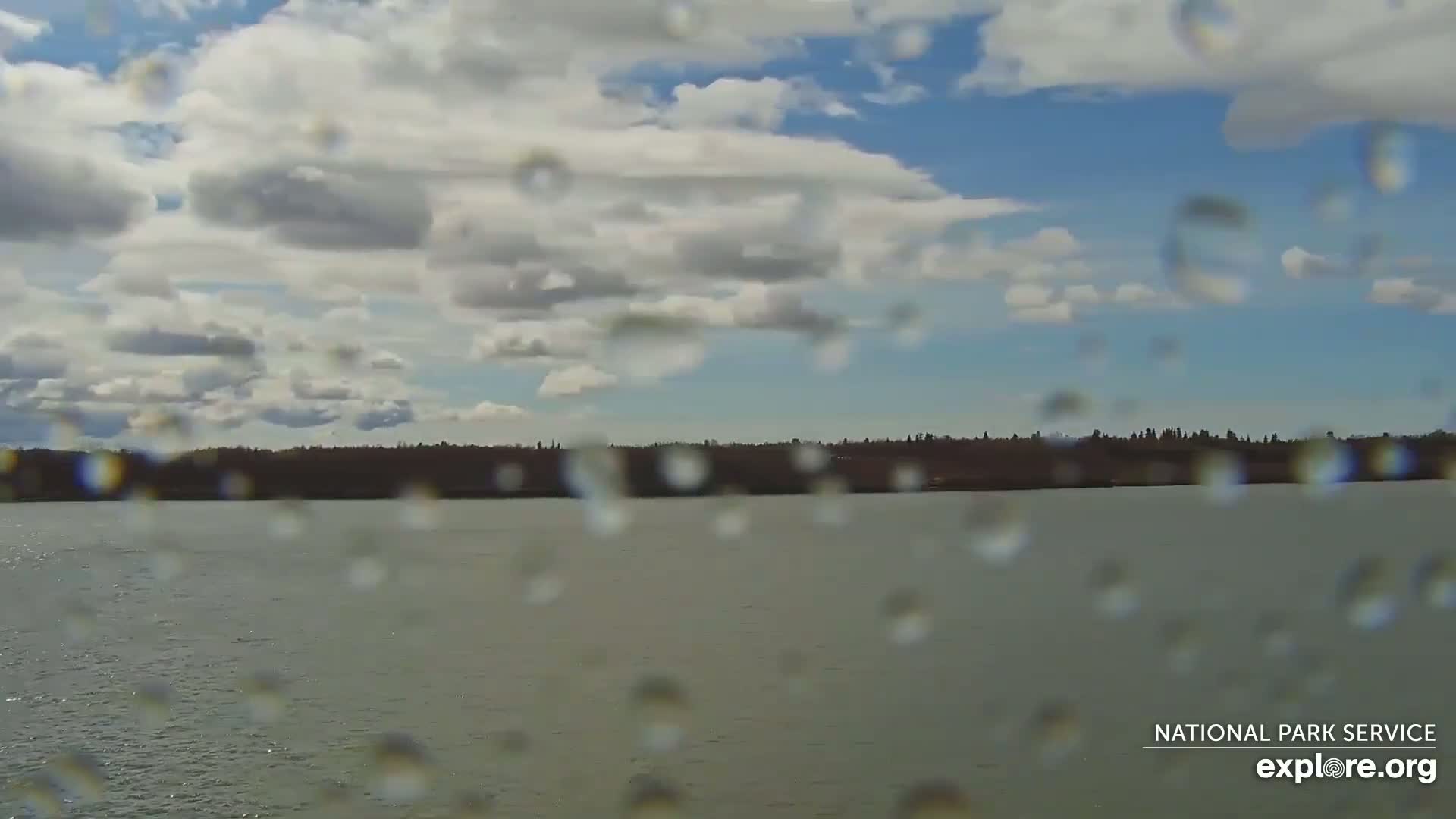 Rain falls on a window overlooking a calm body of water with a distant tree line under a partly cloudy sky.