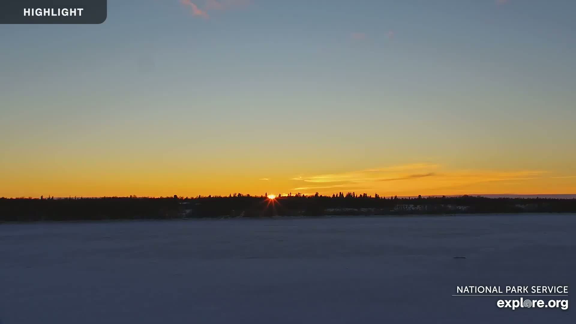 A winter sunset illuminates a snow-covered lake and silhouetted tree line.