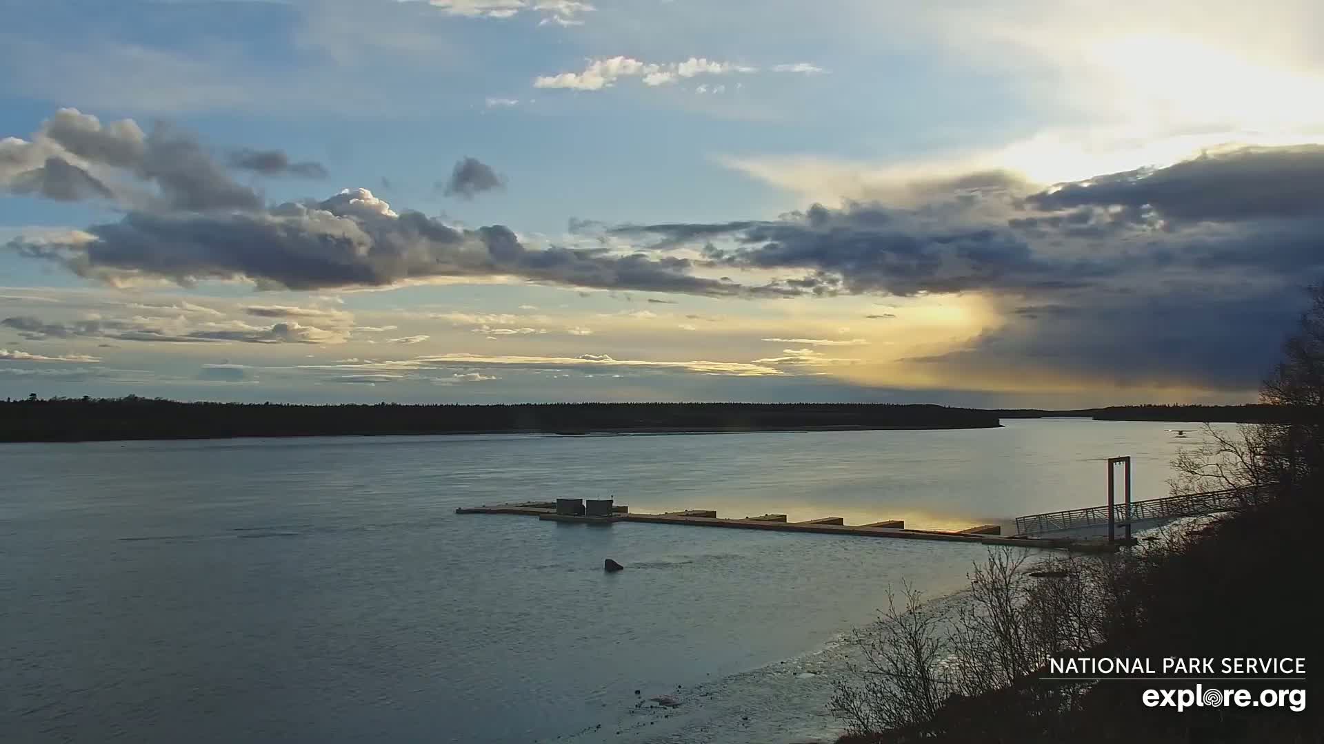 A wide, calm river flows past a small dock under a partly cloudy sky with a sunset breaking through the clouds.