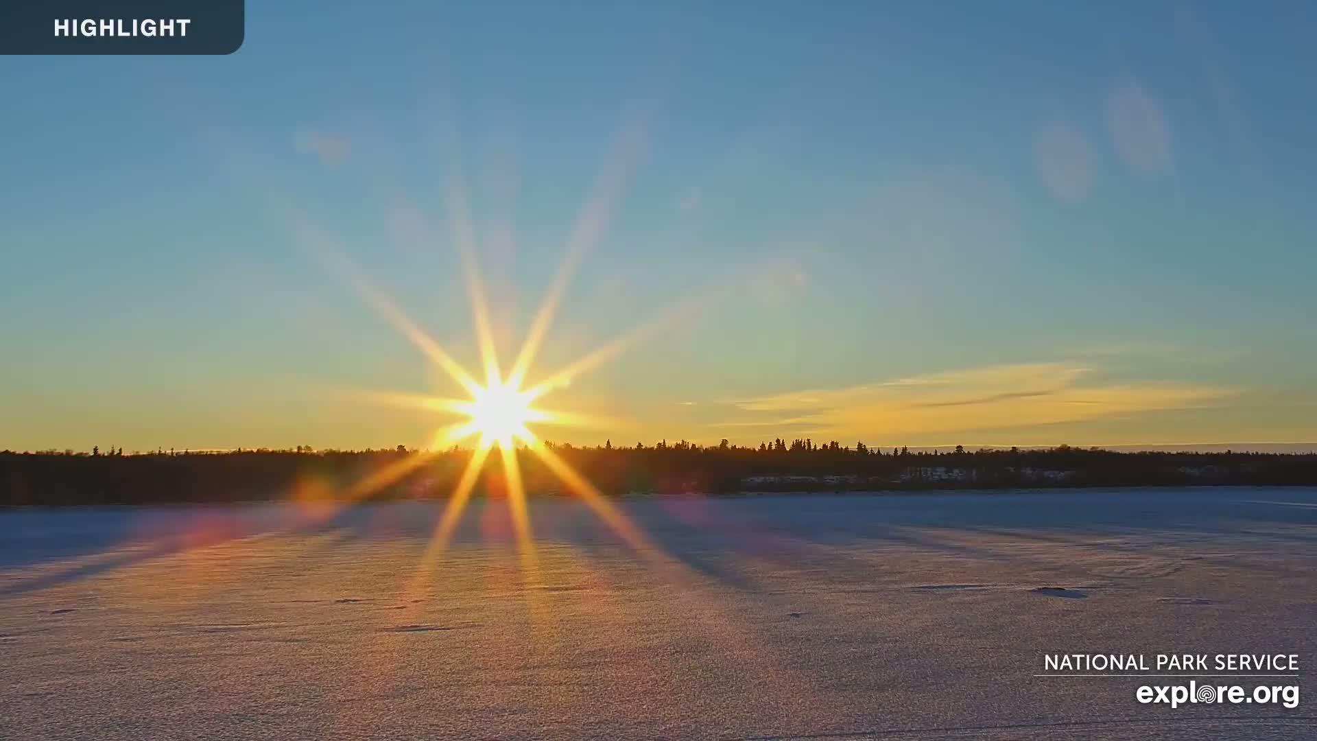 A bright, starburst sun sets over a frozen lake and silhouetted trees.
