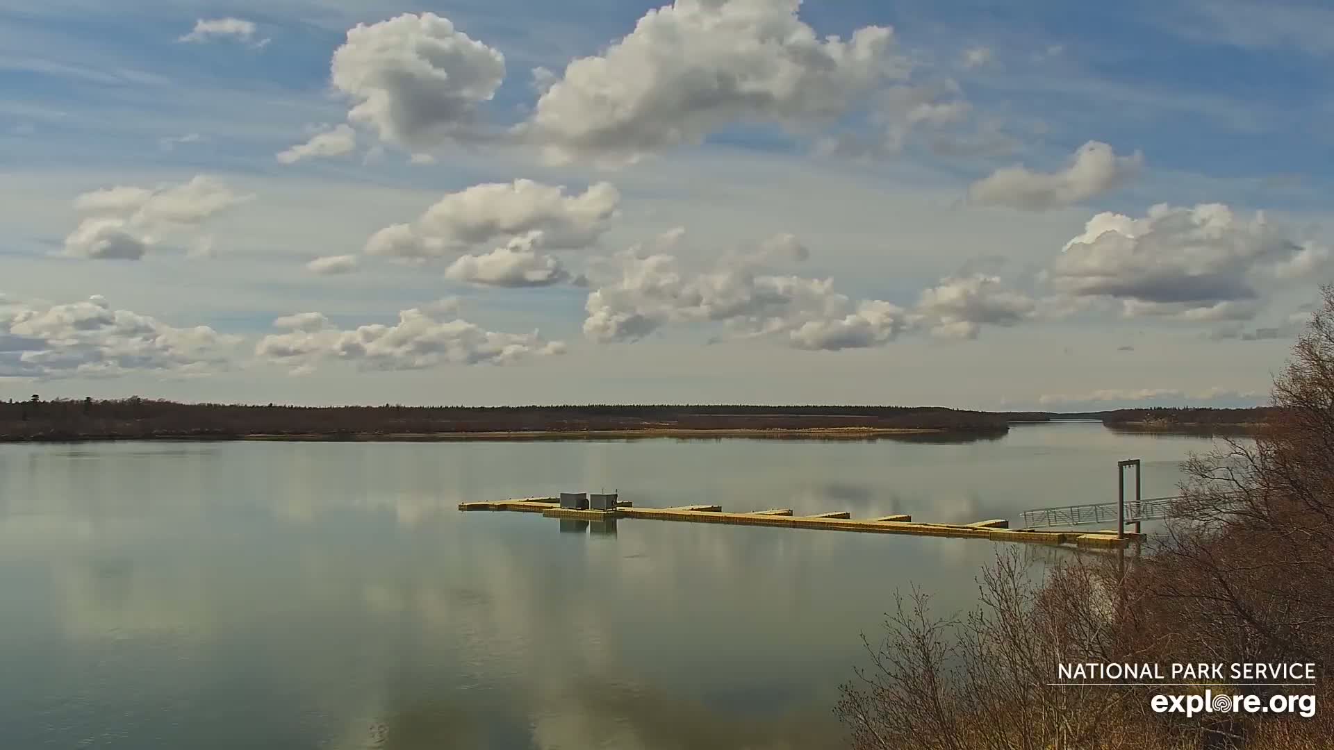 A calm river with a floating dock stretches across the water under a partly cloudy sky.