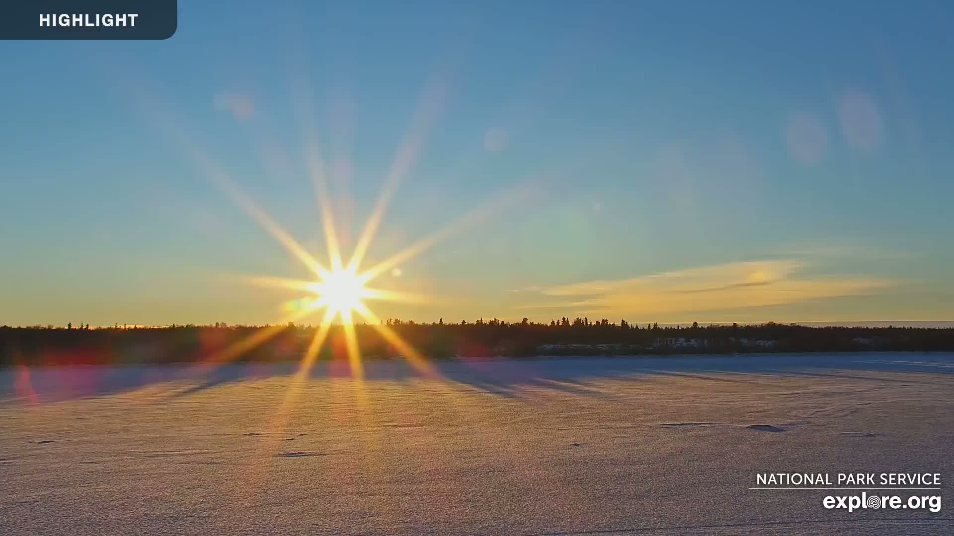 A bright sun sets over a snow-covered frozen lake and a dark treeline.