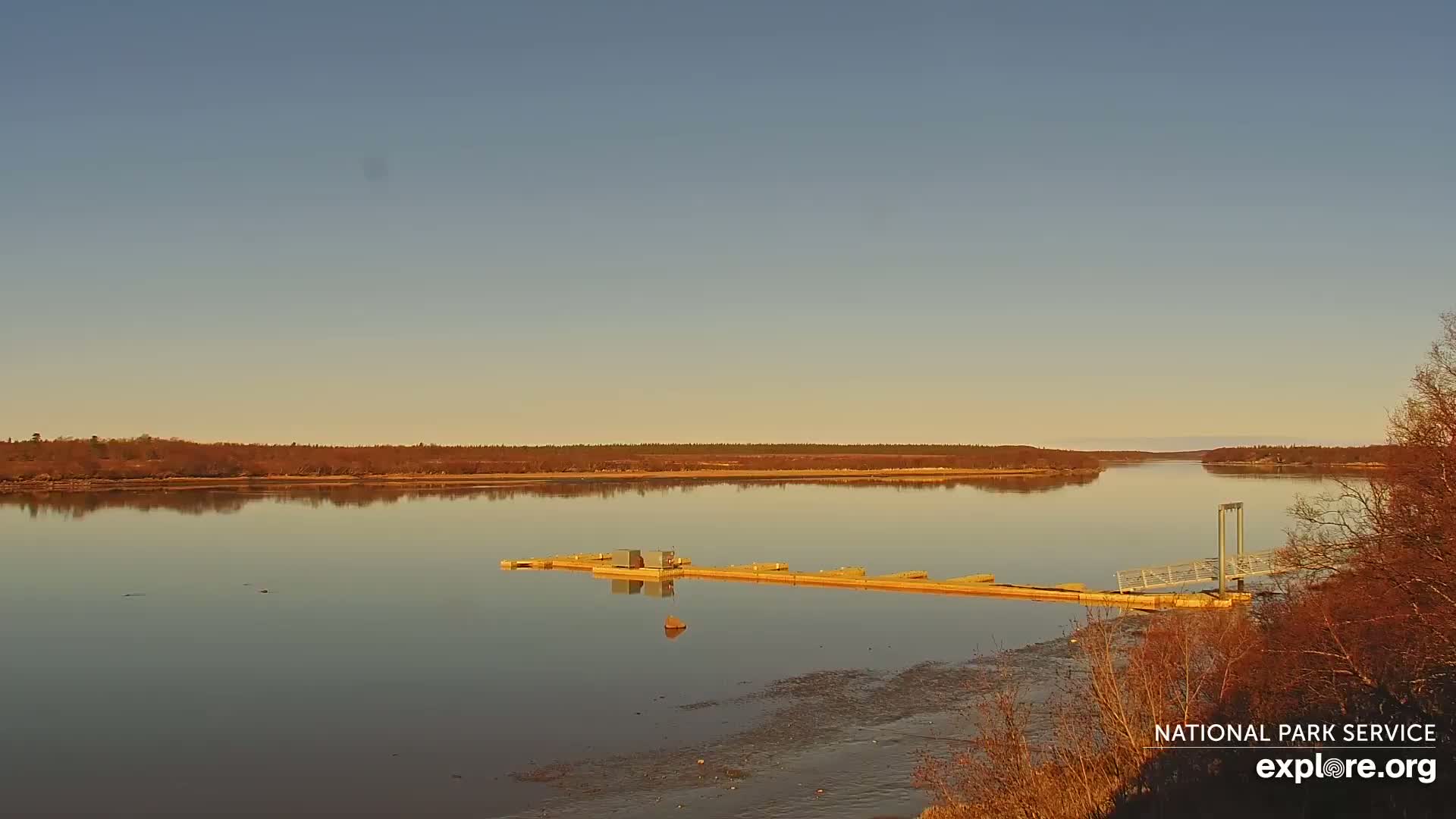 A calm river reflects a pale, clear sky under which a long dock extends into the water, bordered by muted brown trees.