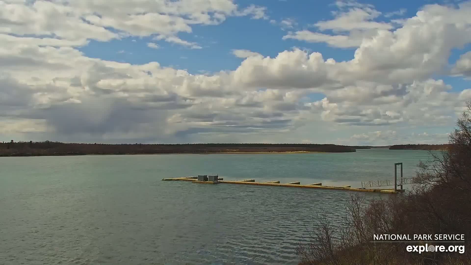 A partly cloudy day reveals a calm body of water with a long dock extending into it, a treeline in the distance.