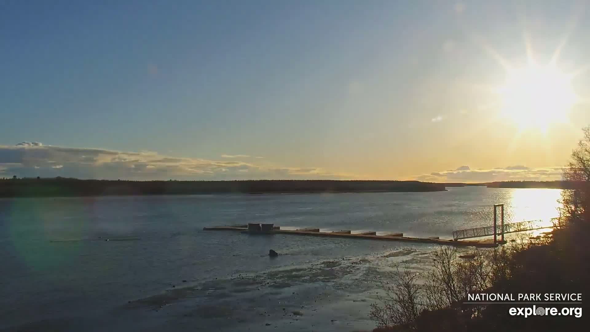 A calm, sunlit river stretches to a tree-lined horizon under a mostly clear sky.