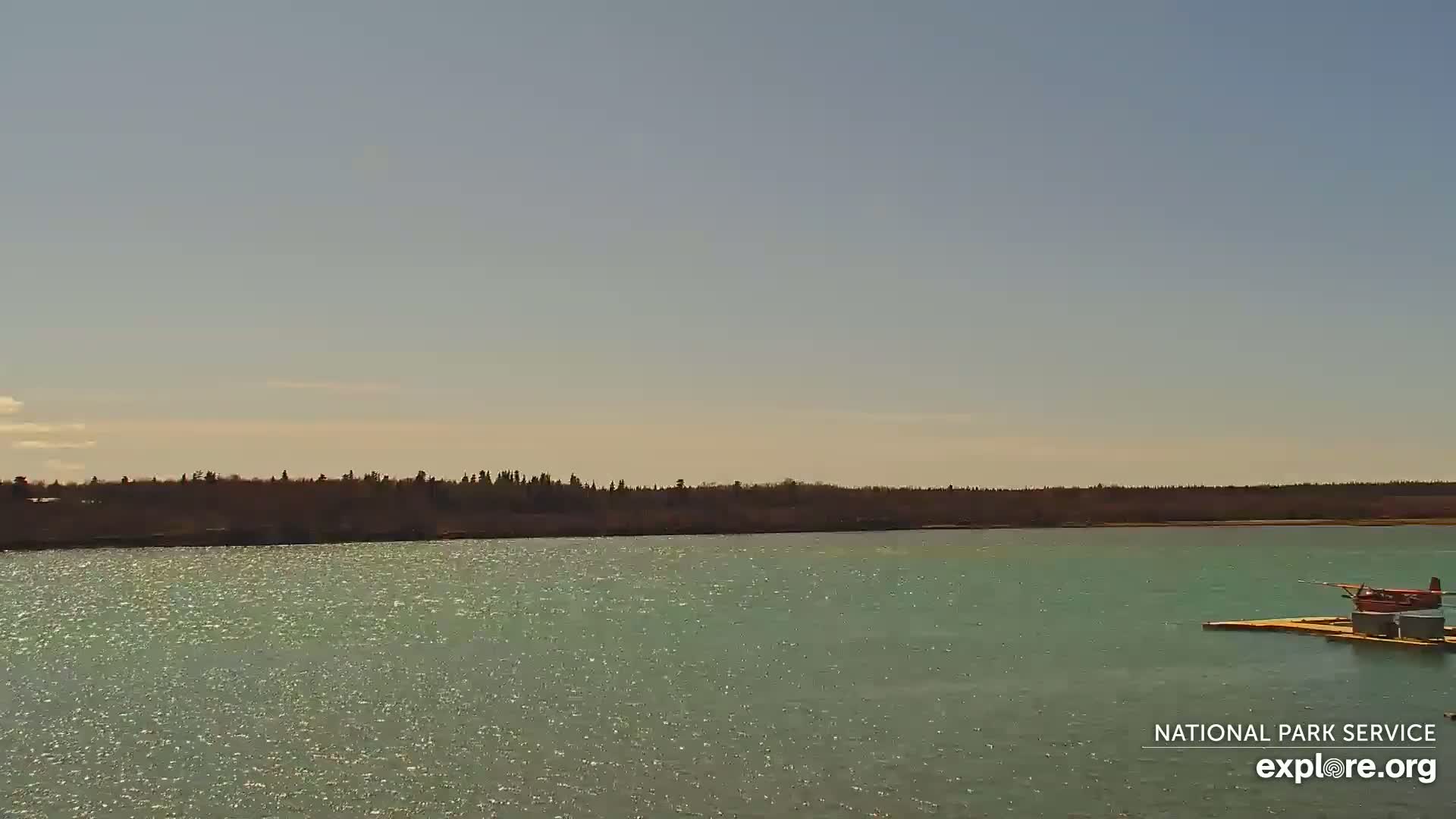 A red seaplane is docked on a platform in a calm, sparkling body of water, with a treeline visible in the distance under a mostly clear sky.