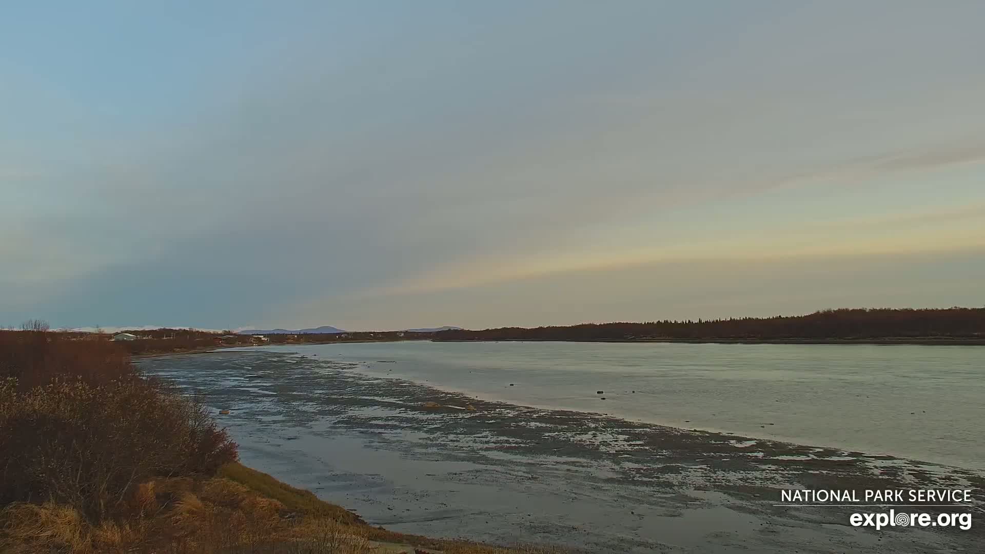 A wide, calm river flows past a flat, muddy shoreline under a pale, mostly clear sky.