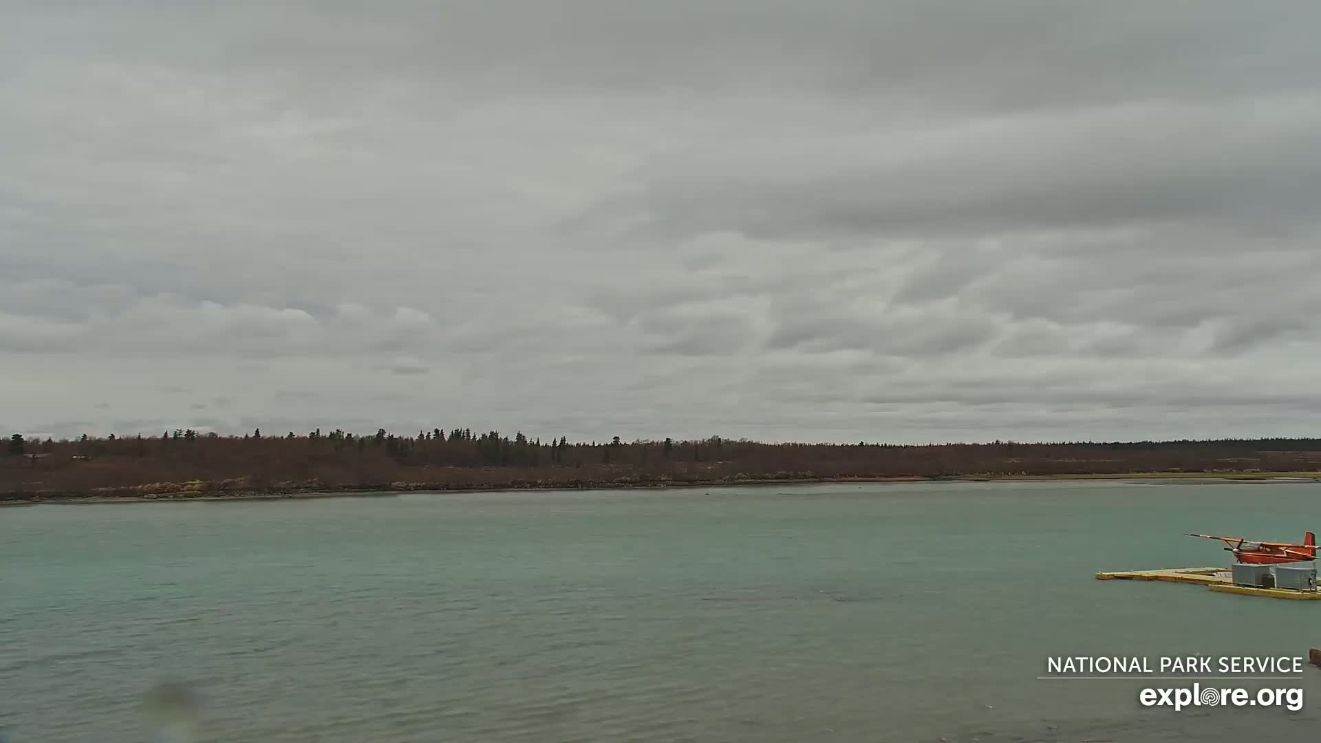 A small, red floatplane is moored on a calm, light-green body of water under a mostly cloudy sky.