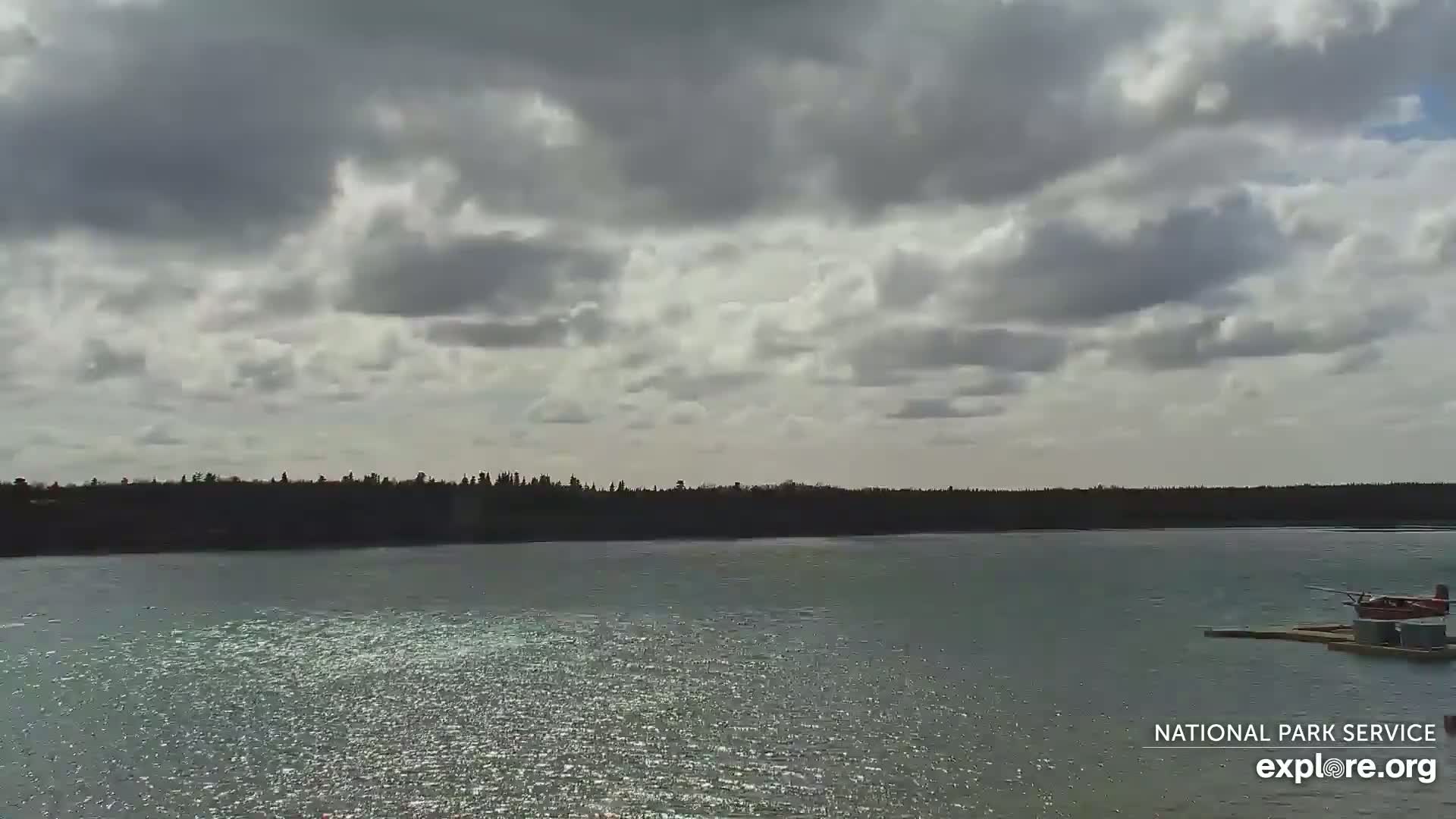 A small red seaplane is docked on a calm body of water under a partly cloudy sky.