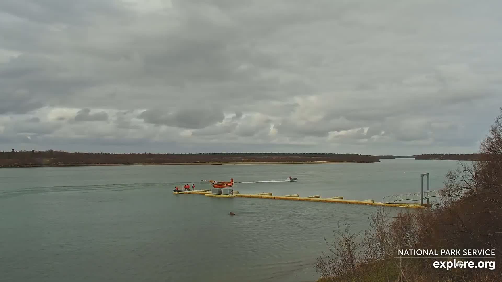 A small orange seaplane is docked at a floating platform on a calm body of water under an overcast sky, with a small motorboat nearby and a shoreline in the distance.