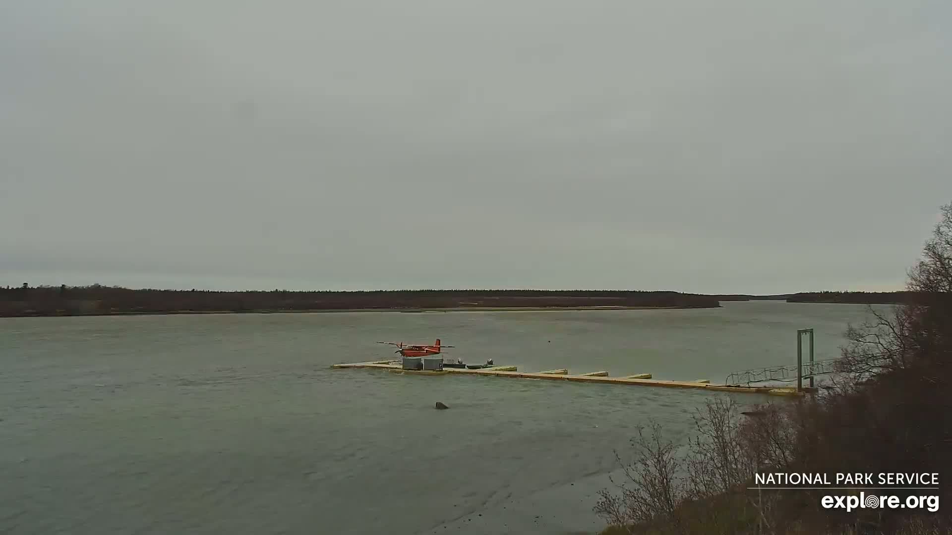 A red seaplane sits on a dock extending into a calm, gray river under an overcast sky.