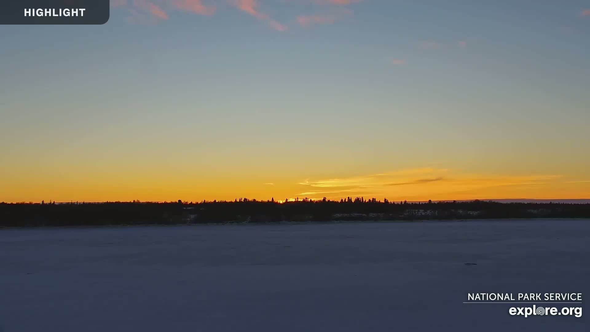 A sunset over a snow-covered expanse of land with a dark treeline in the distance.