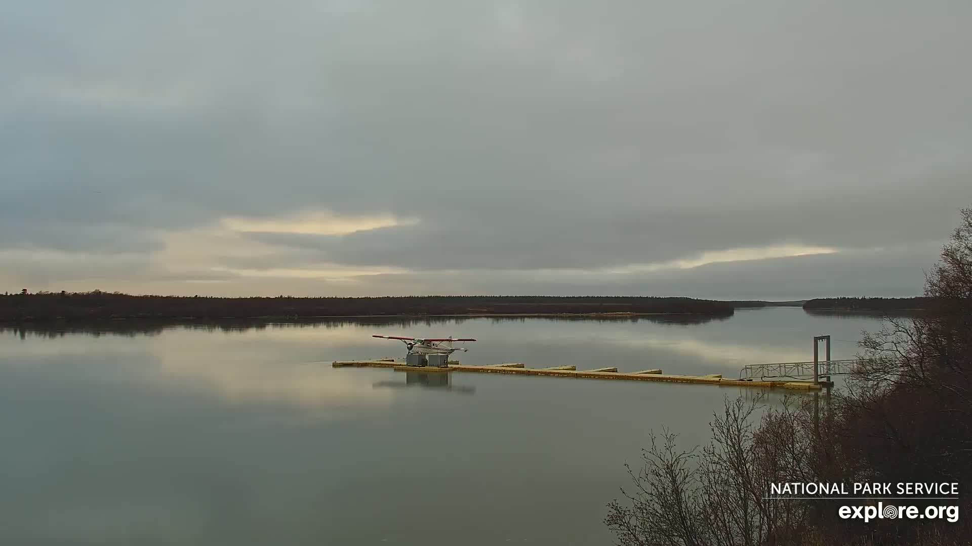 A seaplane rests on a dock in a calm river under an overcast sky.