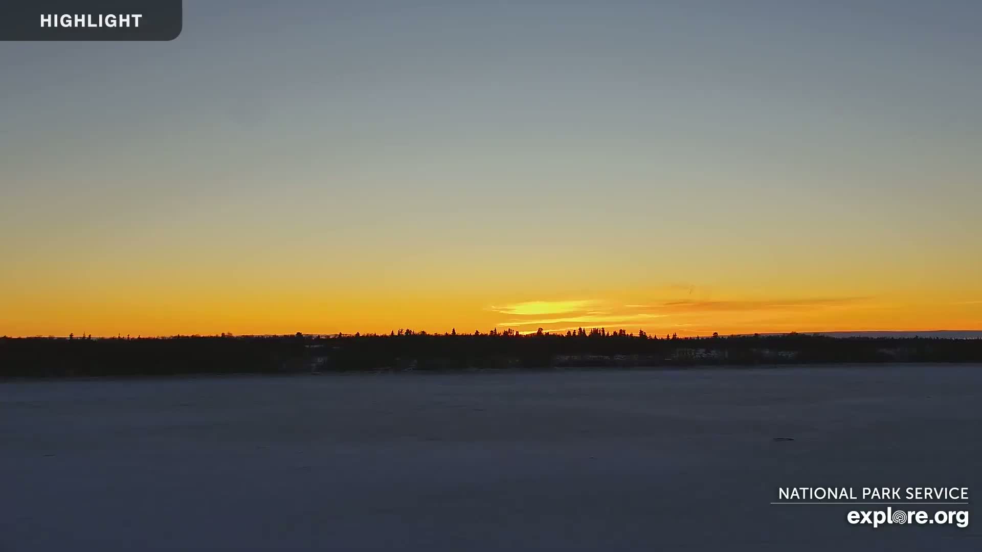 A golden sunset illuminates a snow-covered expanse of land and water, with a dark line of trees silhouetted against the horizon.