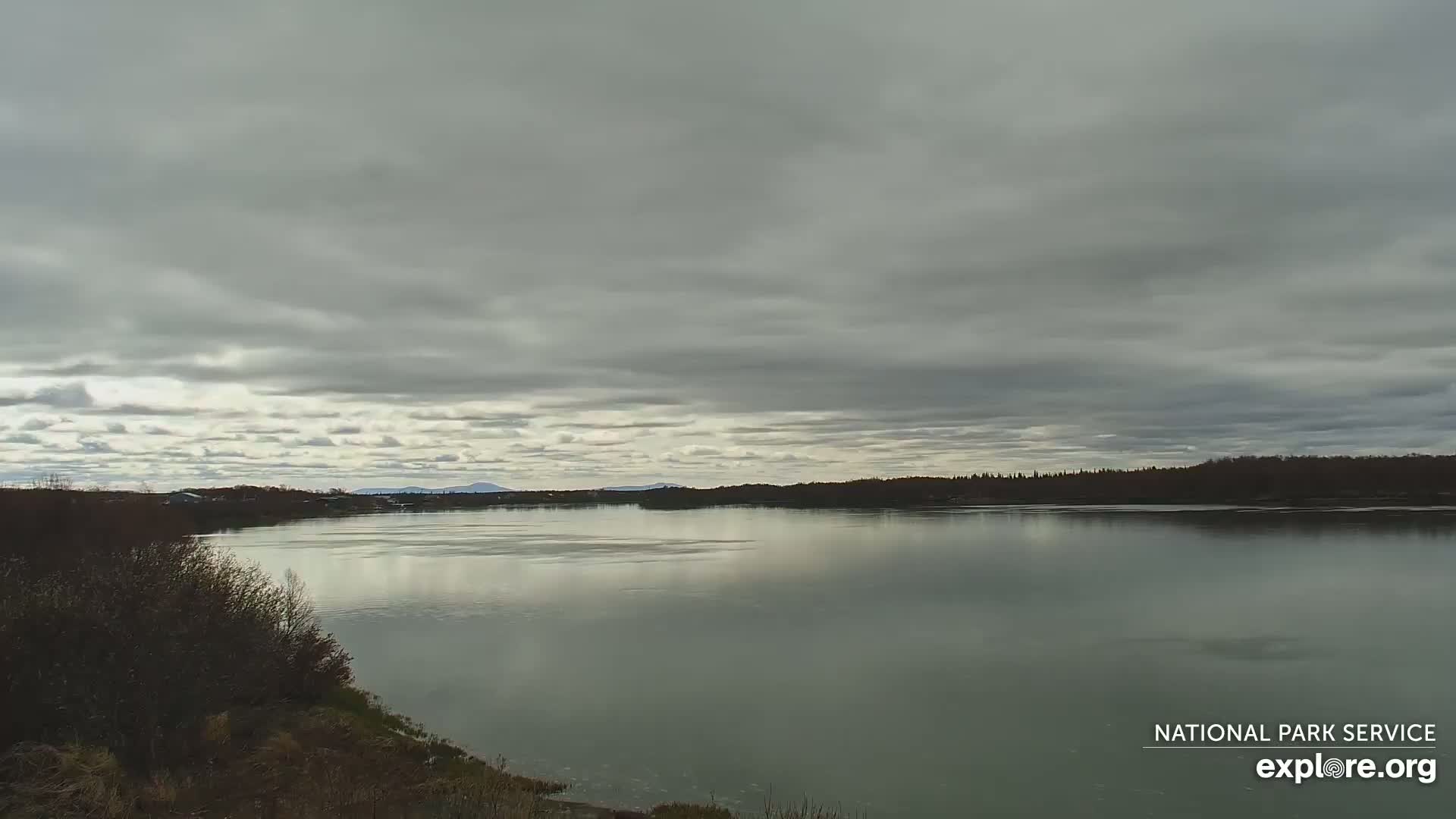 A wide, calm river reflects a mostly cloudy sky under which a distant mountain range is barely visible.