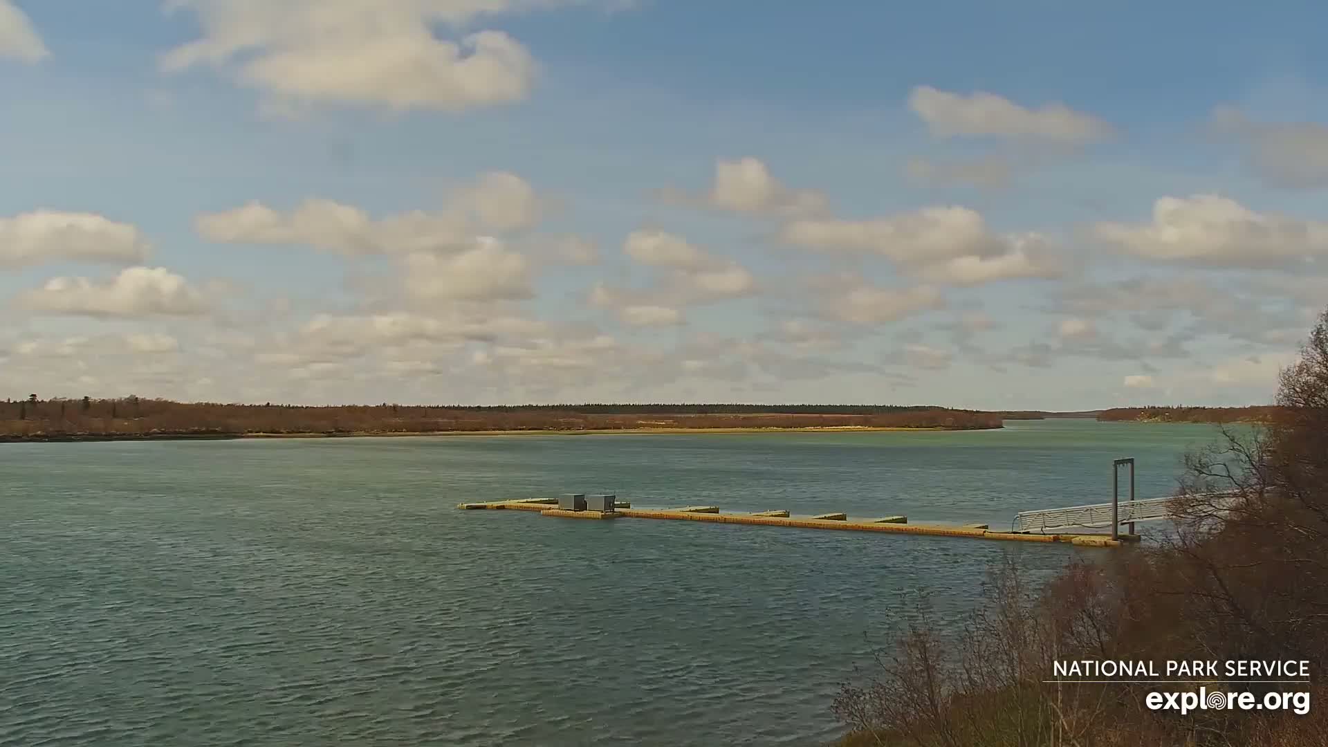 A long dock extends into a calm body of water under a partly cloudy sky, with bare trees visible in the foreground and a treeline across the water in the background.