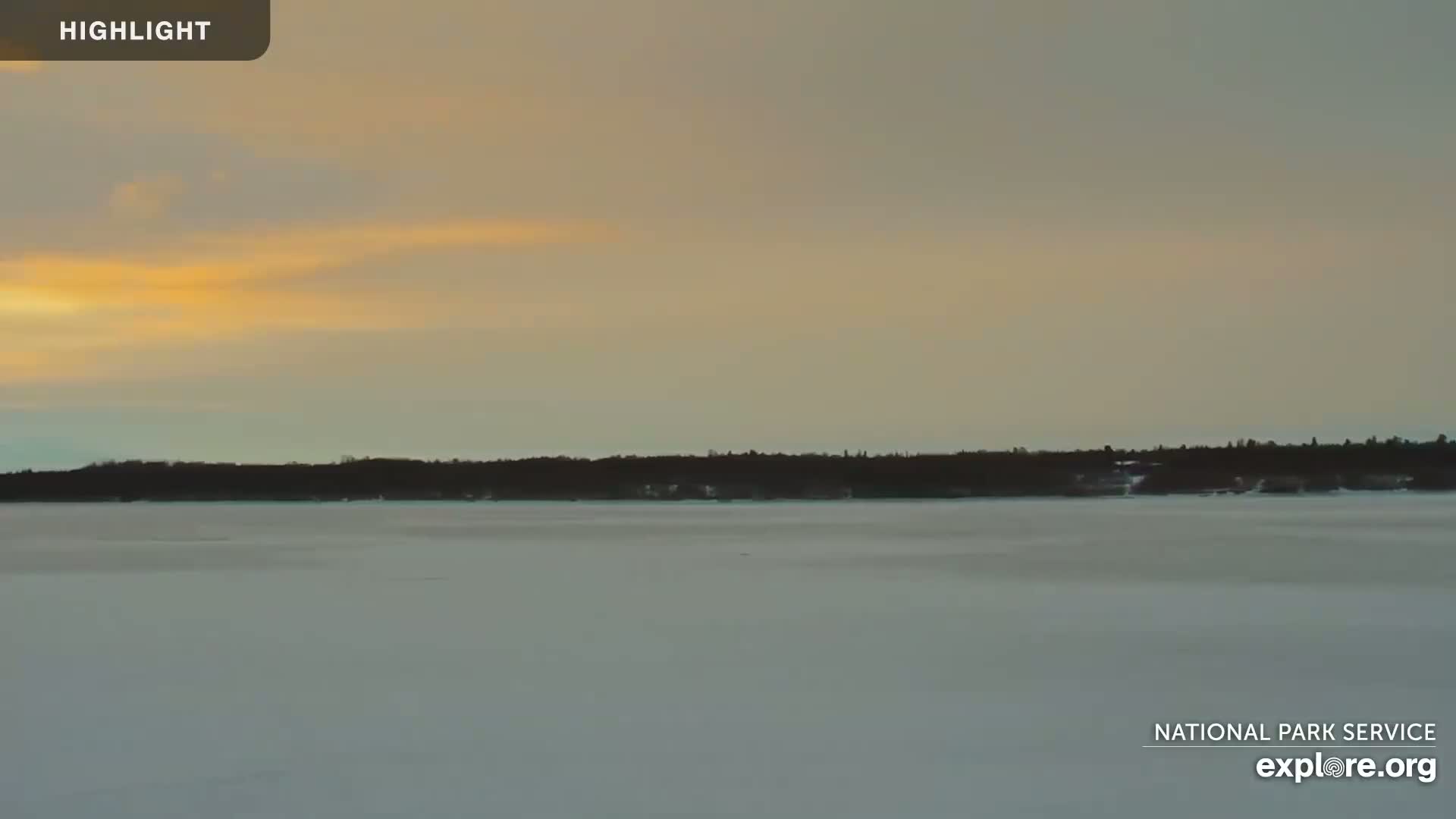 A frozen lake stretches before a dark treeline under a pale, sunset sky.