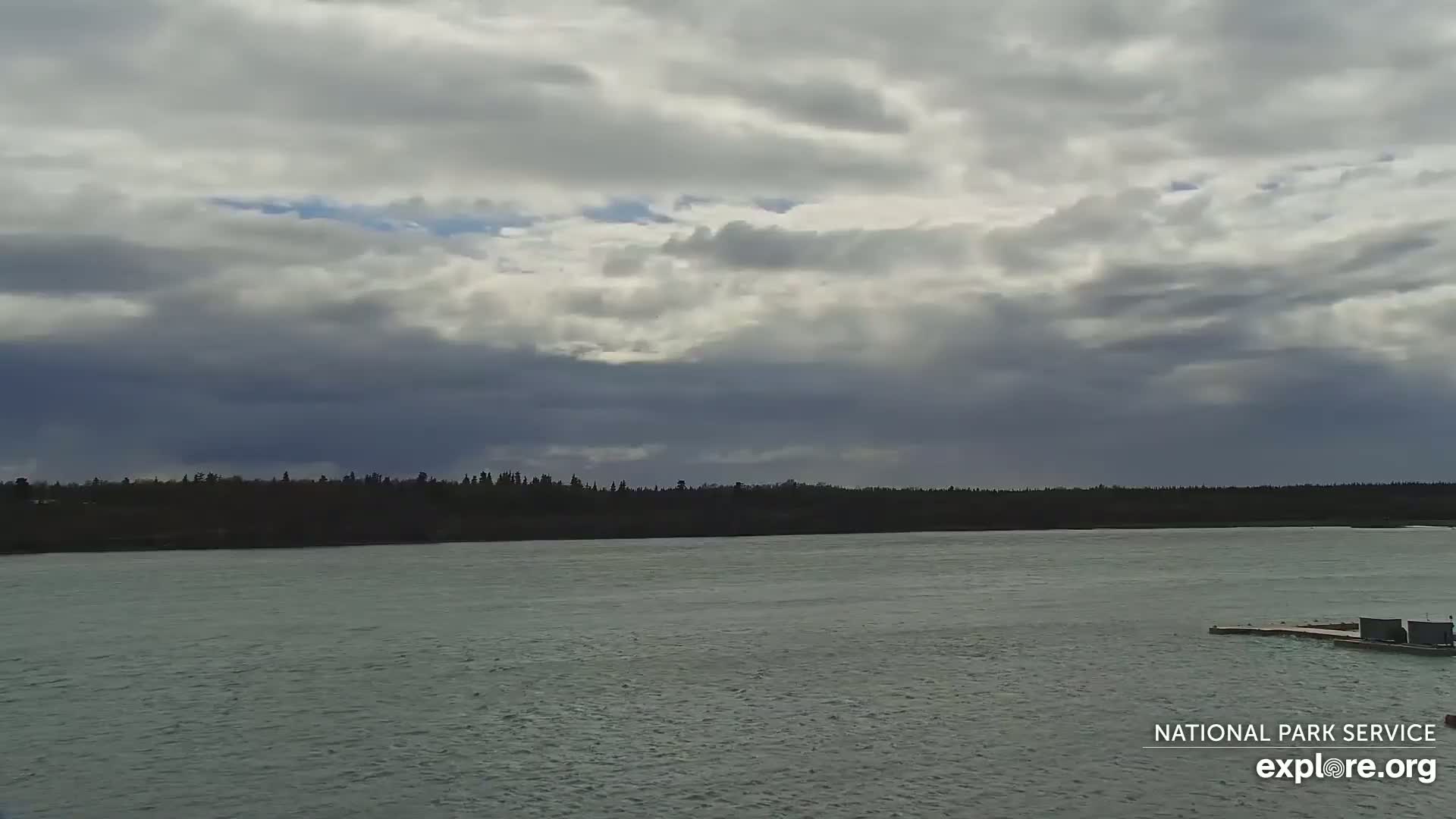 A wide body of relatively calm water stretches out under a mostly cloudy sky, with a dark line of trees visible on the far shore.