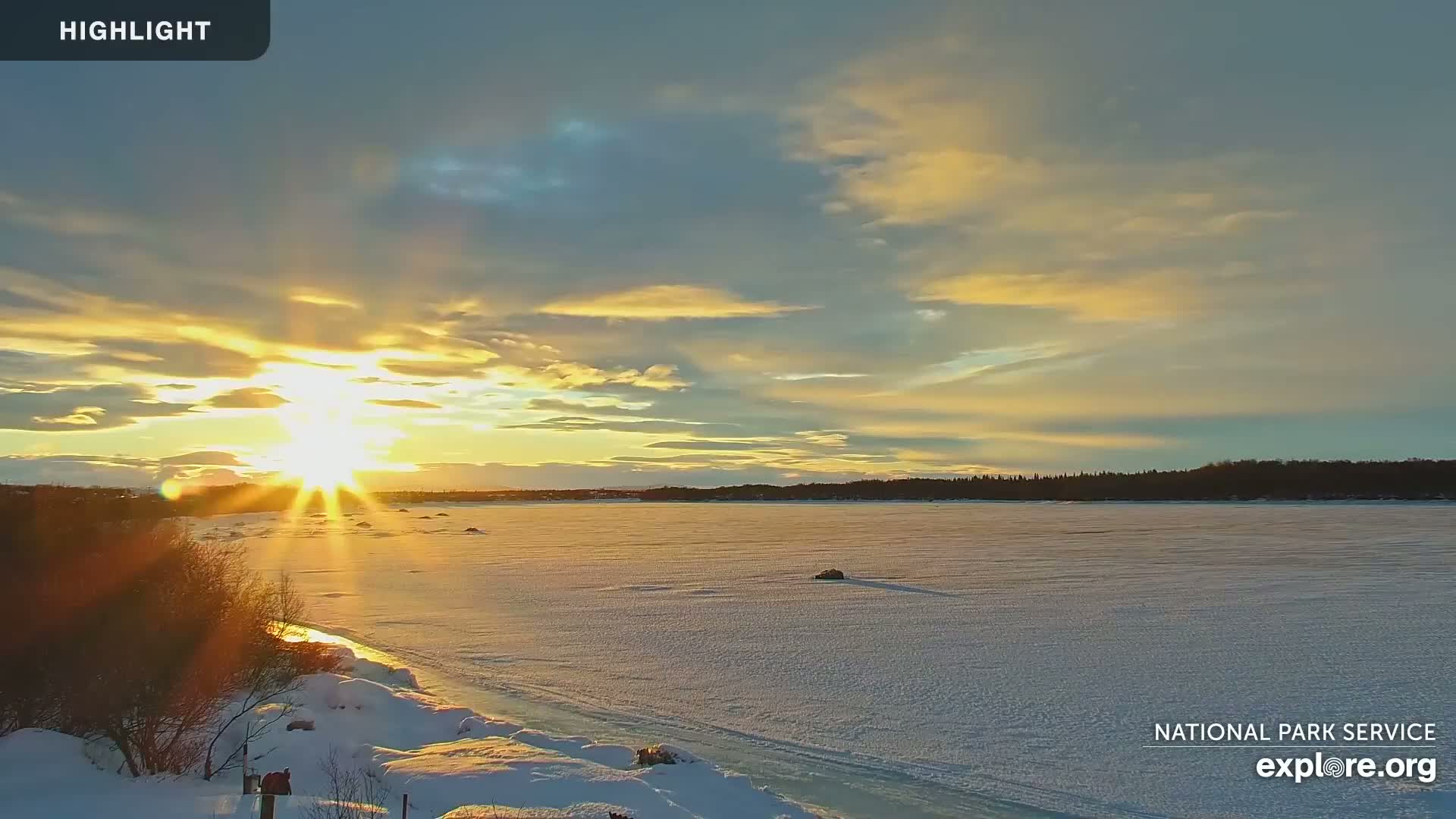 A bright sunset casts a golden glow over a snow-covered frozen lake and shoreline, with a few dark trees in the distance.