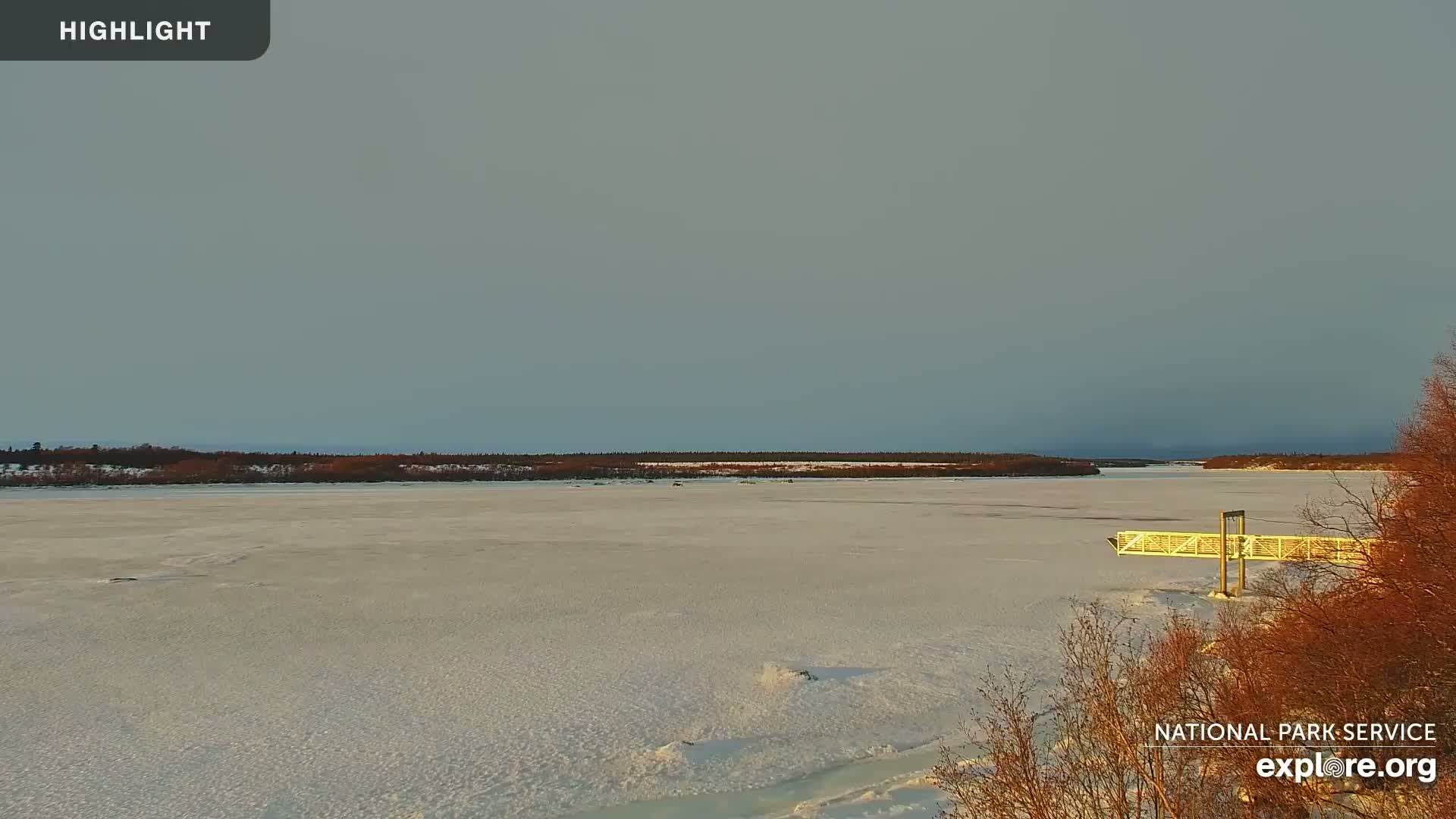 A wide, snow-covered frozen river stretches out under a pale, overcast sky, with a small dock visible near the bottom right and a treeline on the horizon.