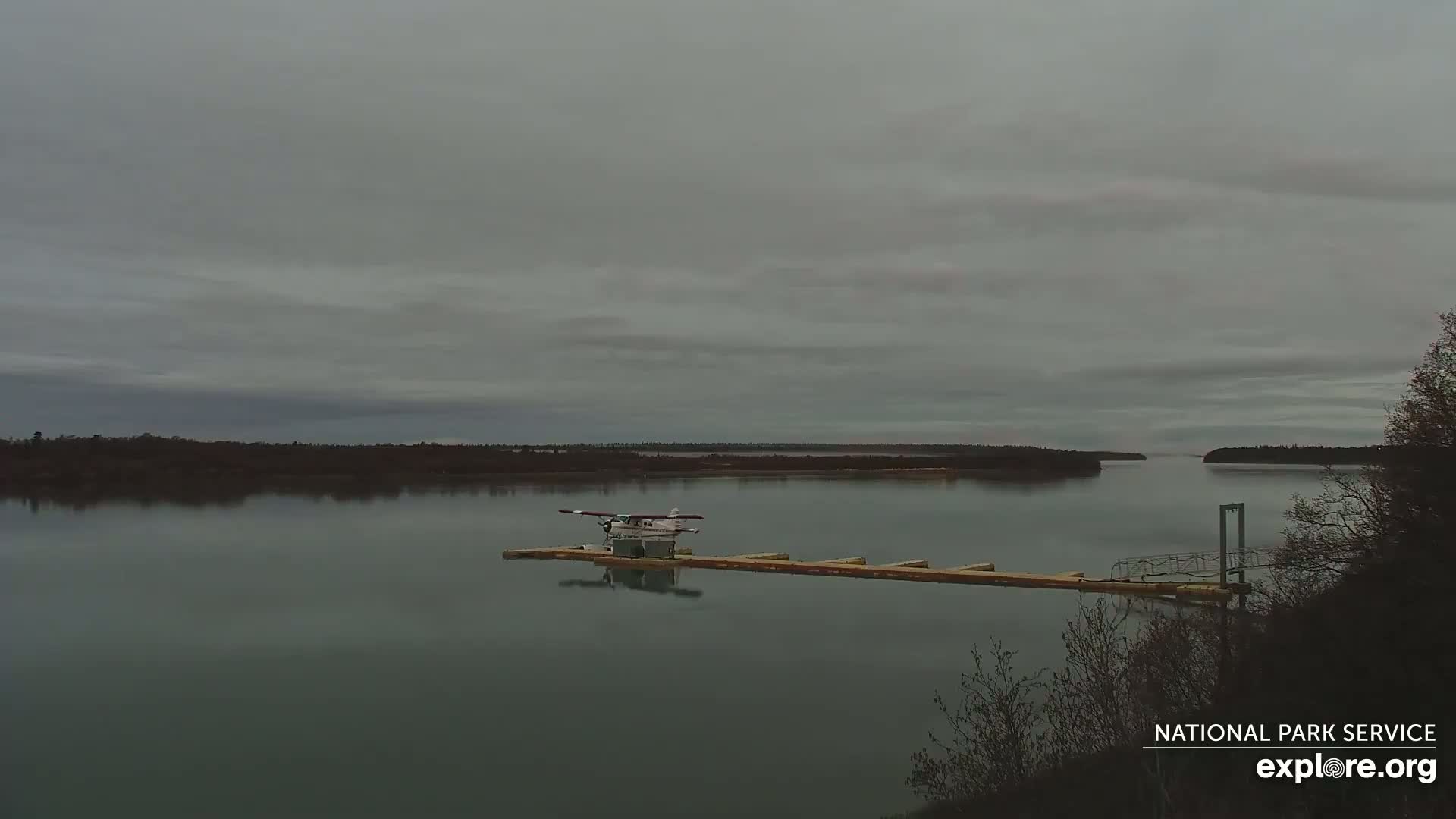 A seaplane is docked at a long, narrow pier on a calm river under an overcast sky.