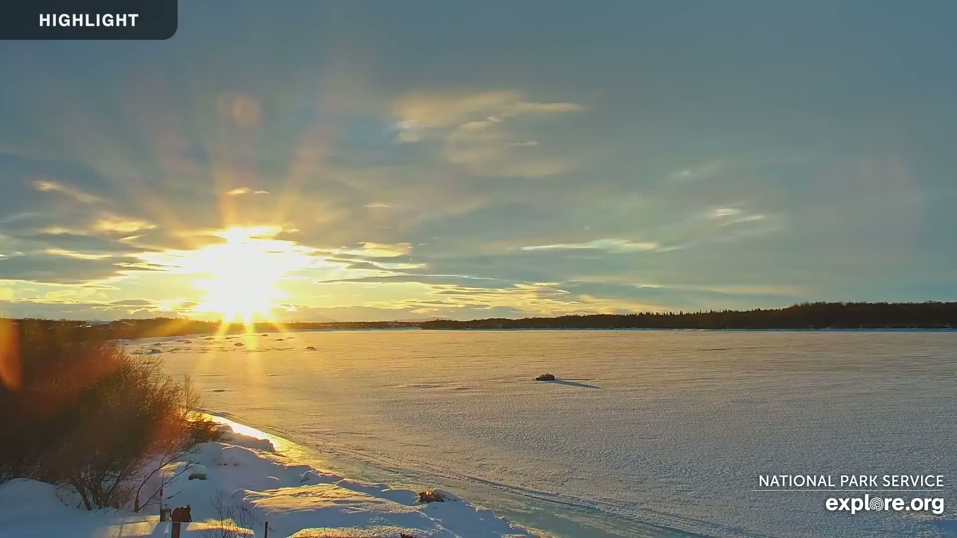 A frozen lake stretches to a tree line under a bright, setting sun with a mostly clear sky.