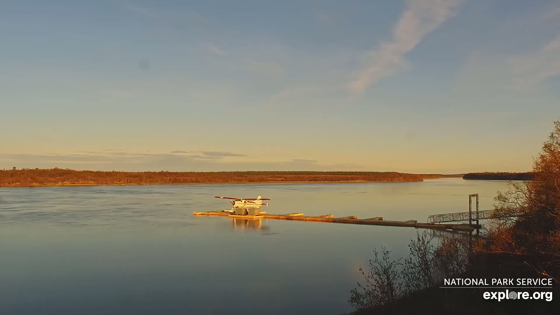 A seaplane is docked at a small pier on a calm river under a clear, pale-orange sunset sky.