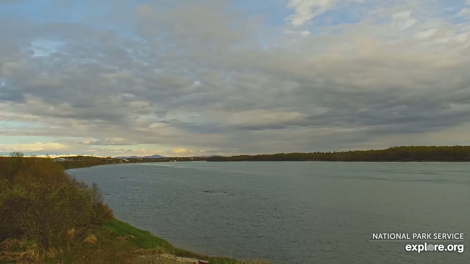 A wide, calm river flows under a mostly cloudy sky, with a shoreline of trees and low hills in the distance.