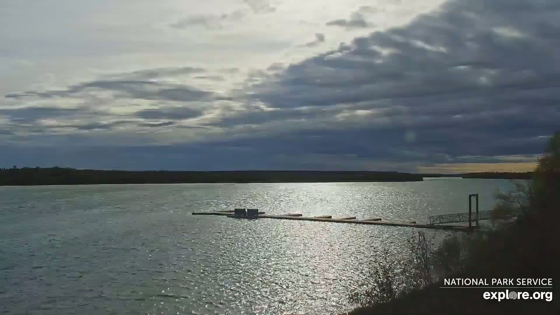 A mostly cloudy sky hangs over a calm river with a dock extending into the water.