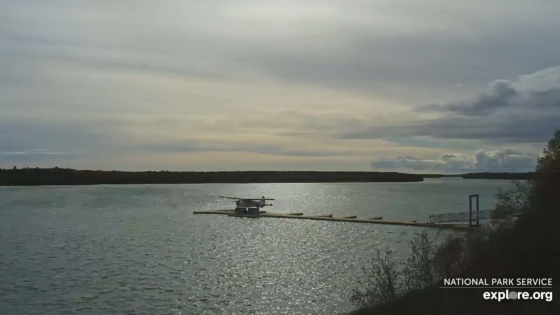 A seaplane is docked at a small pier on a calm lake under a partly cloudy sky.
