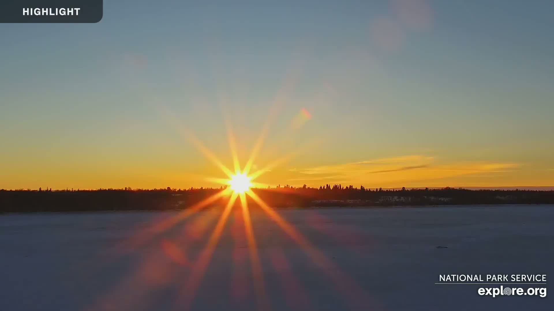 A bright, starburst sun sets over a snow-covered lake and a dark treeline.