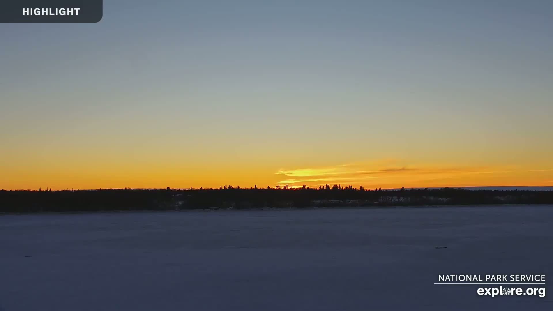 A sunset casts a warm orange glow on a snow-covered lake and silhouetted tree line.