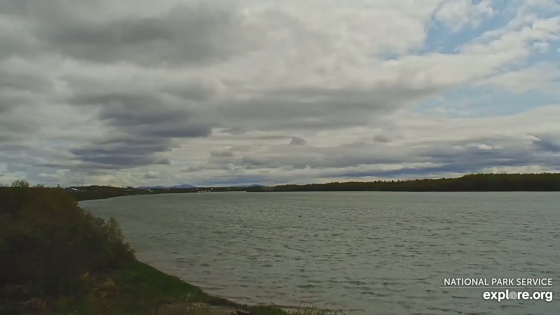 A wide, calm river stretches to a treeline under a mostly cloudy sky.