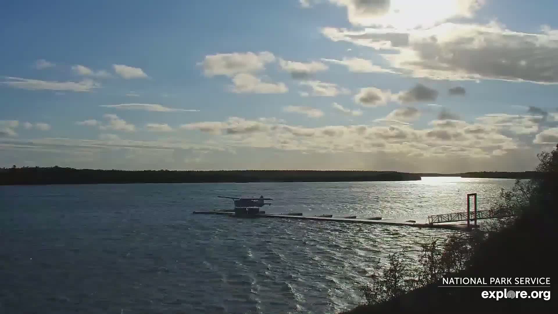 A seaplane is docked at a floating platform on a calm, partly cloudy day on a wide body of water.