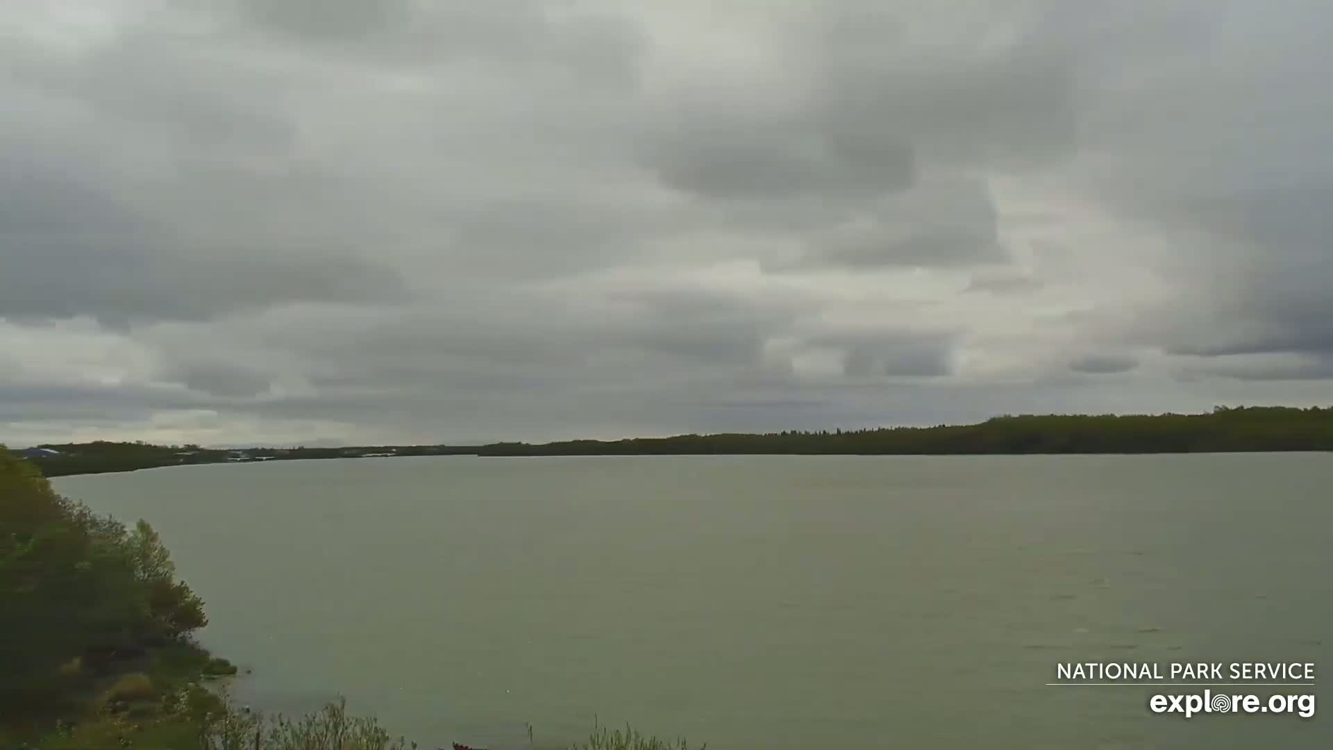 A wide, calm body of water stretches to a tree-lined shore under a cloudy sky.