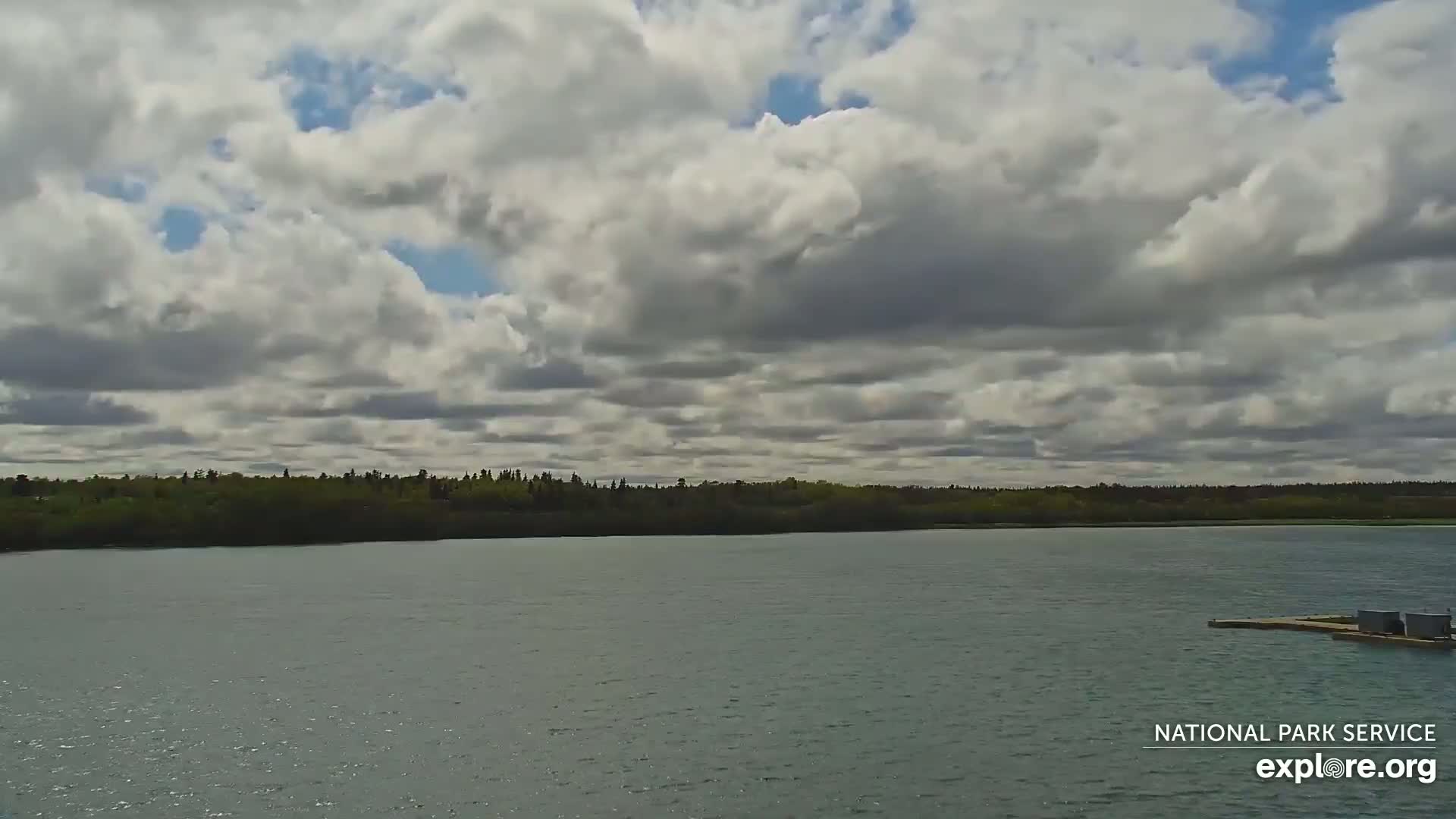 A mostly cloudy sky overlooks a calm body of water with a treeline in the distance and a small dock to the right.