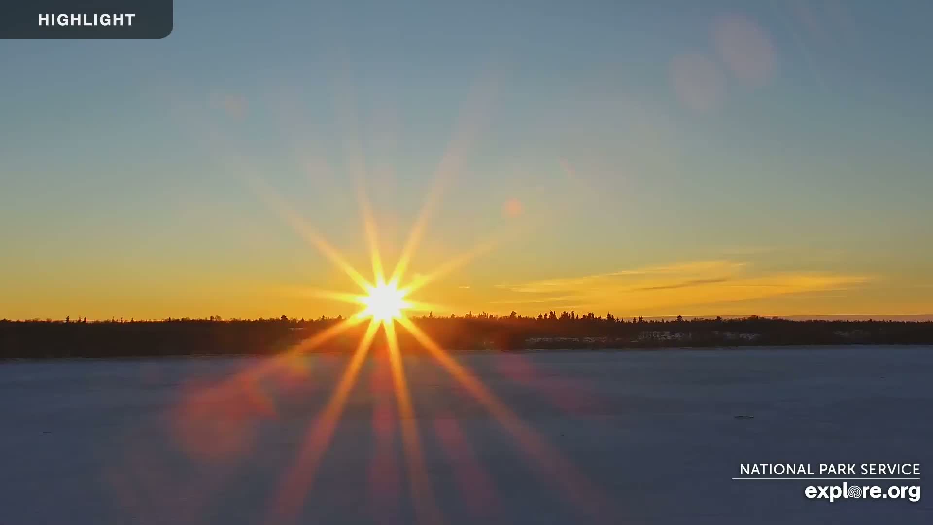 A bright sun with lens flare sets over a snow-covered lake and dark tree line, under a clear, orange and blue sky.