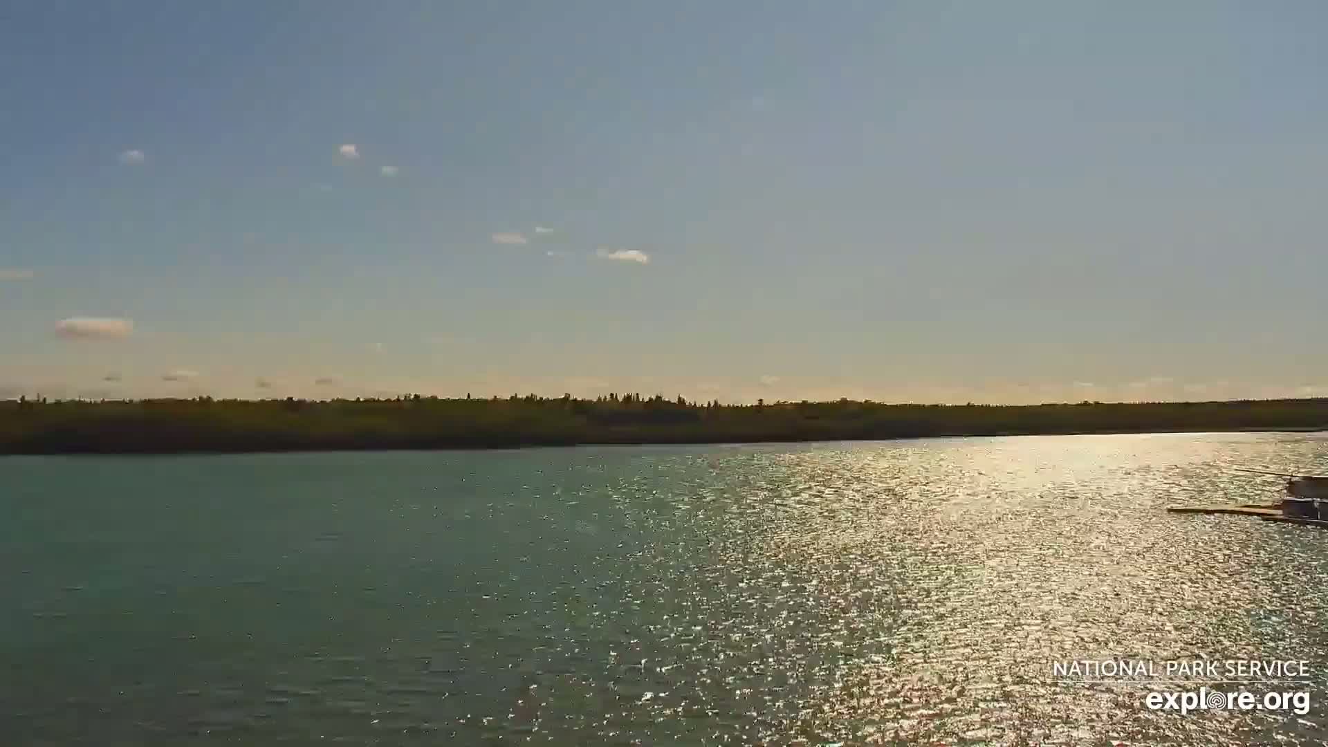 A calm, sunlit river reflects the light, with a treeline visible in the distance under a mostly clear sky.