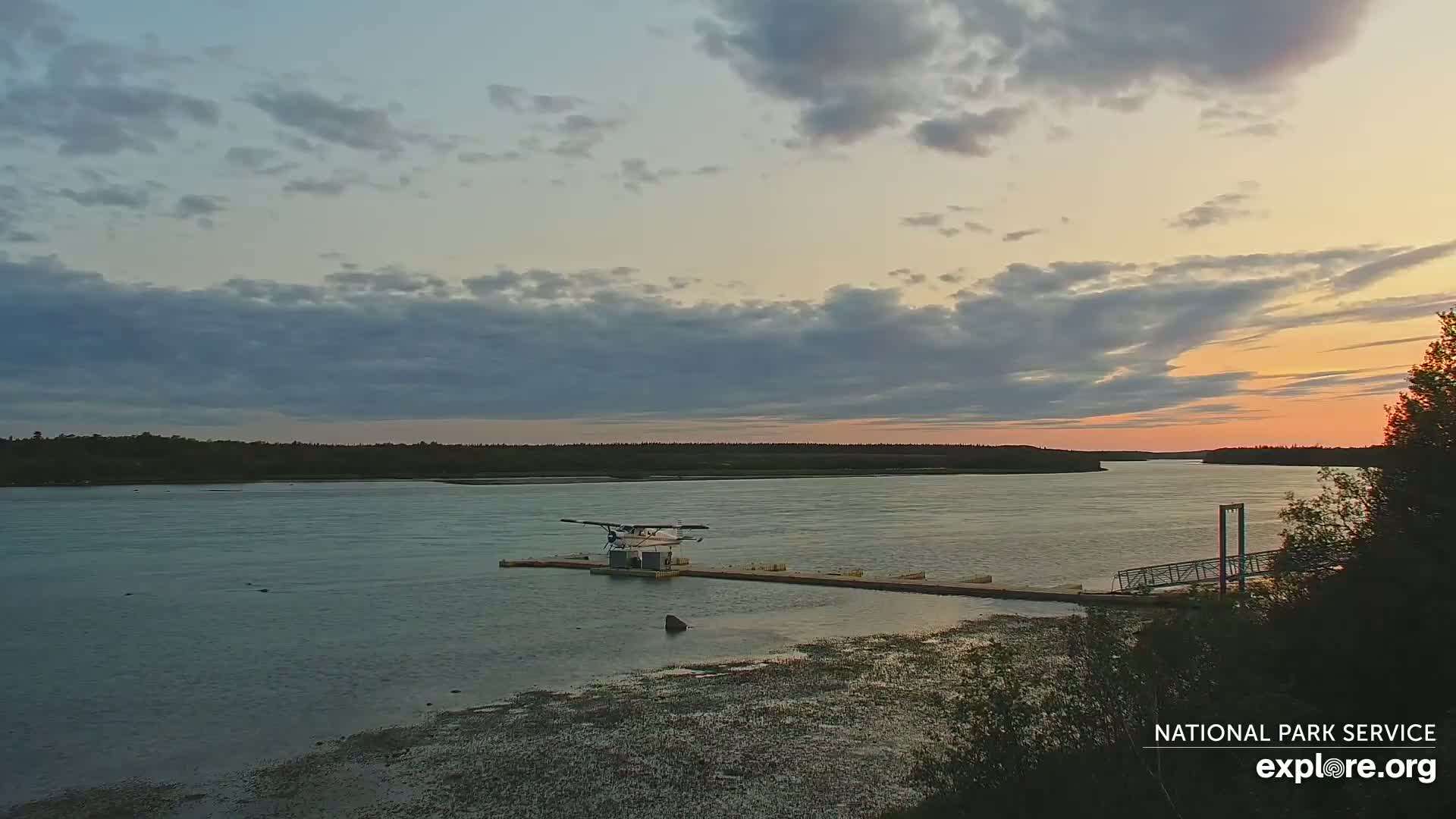 A small seaplane is docked at a small pier on a calm river under a partly cloudy sunset sky.