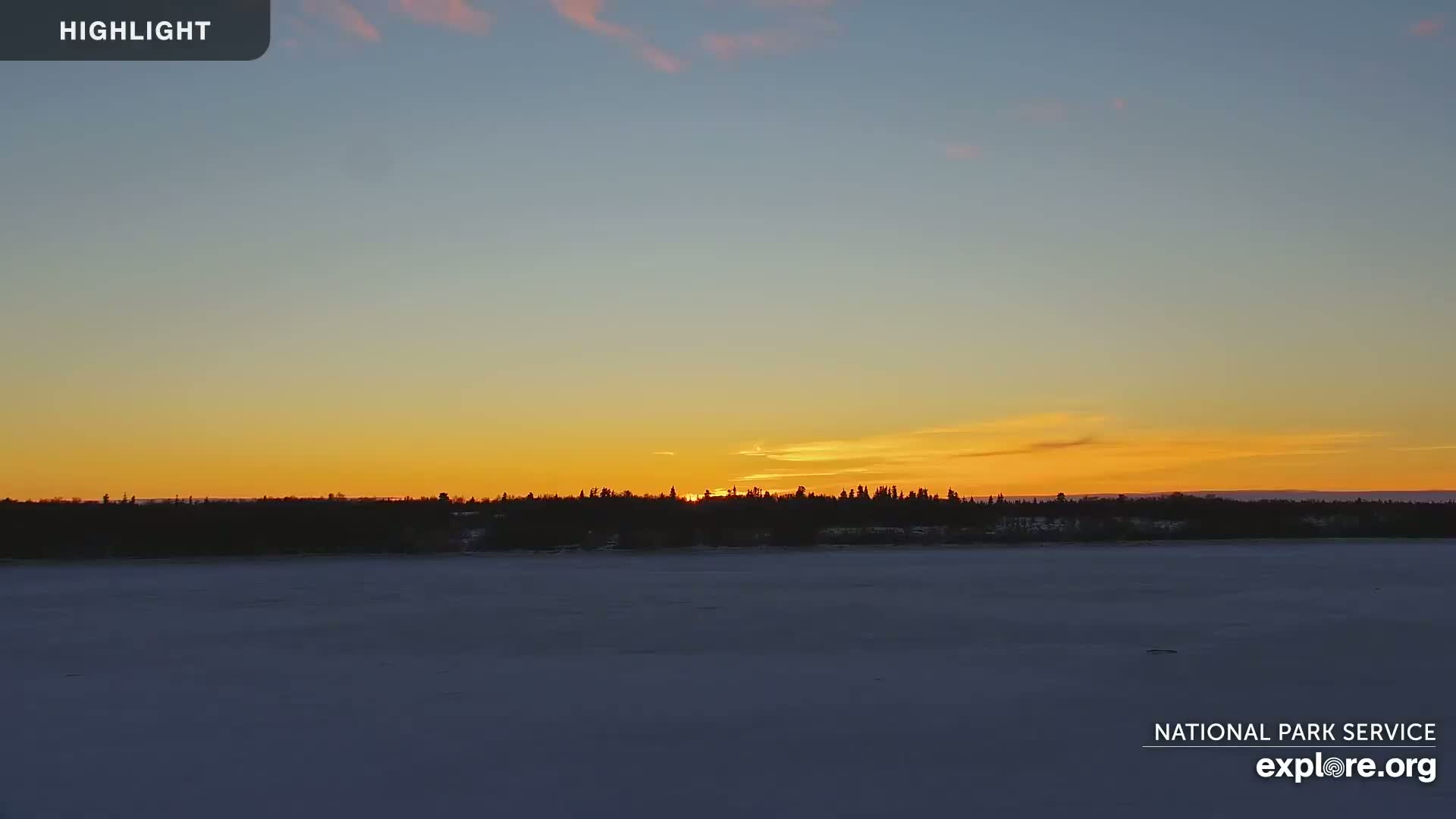 A sunset casts a warm orange glow over a snow-covered frozen lake and a dark silhouetted treeline.