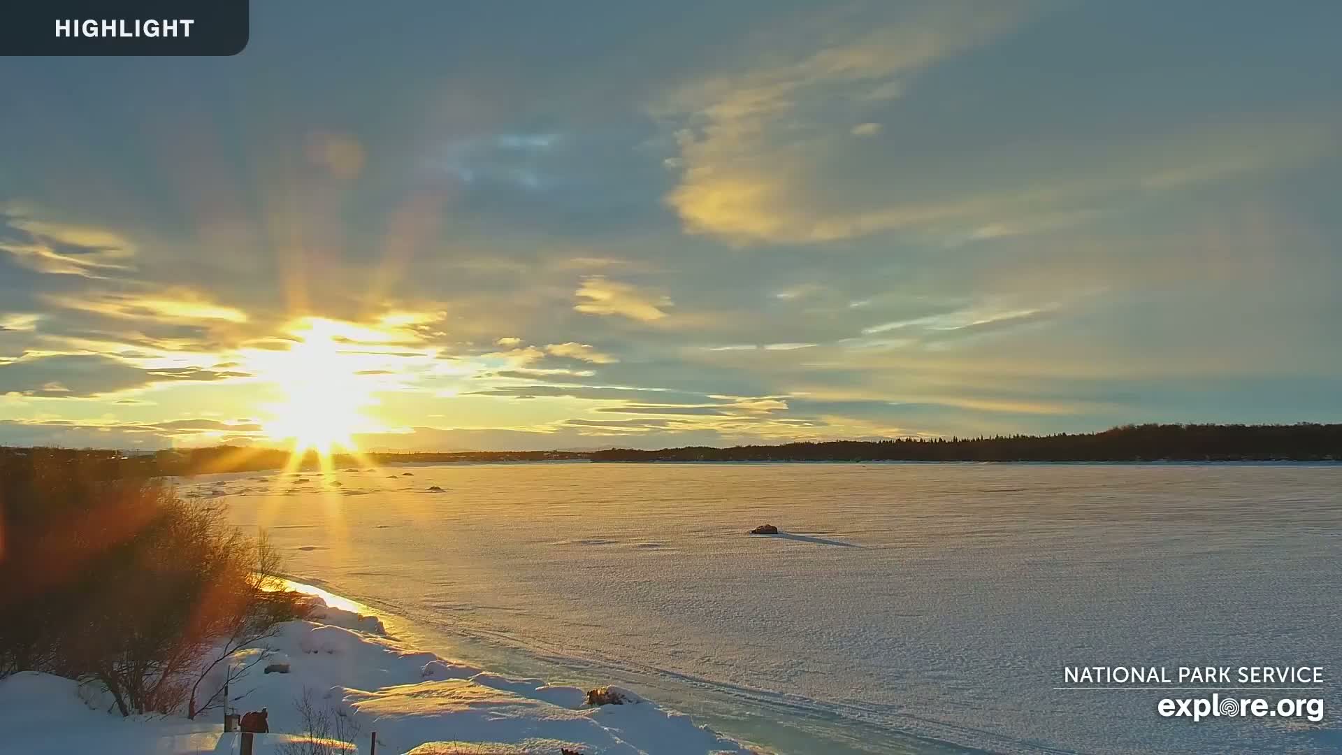 A bright, low sun sets over a frozen, snow-covered lake with a treeline in the distance.